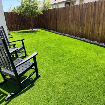 Photo of a sunny backyard with a bright green lawn, brown wooden fencing, and 2 black Adirondack style rocking chairs on the grass, showing a positive answer to the question "is artificial turf worth it"