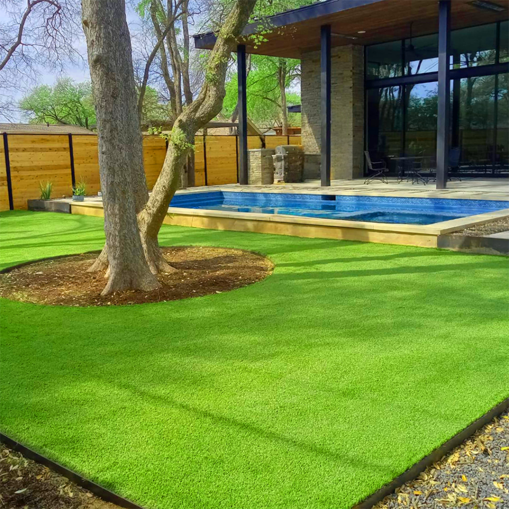 Section of a yard with artificial turf around a pool and a large tree shading the space. A modern covered porch attached to the house is behind the pool.