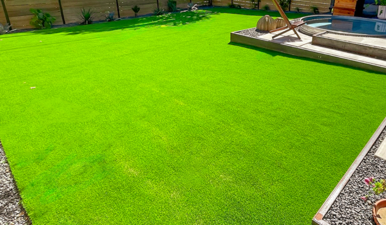 Sunny residential backyard of a customer who might ask "Does artificial turf get hot?" There is a small in-ground hot tub in the background, a raised garden bed in the foreground, and the yard is fenced in with some plants decorating the edge of the yard.