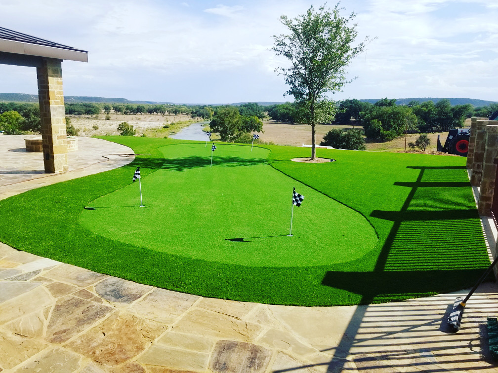 A large backyard putting green with a wide view of green hills. The green has several holes, each marked with a black and white checkered flag.