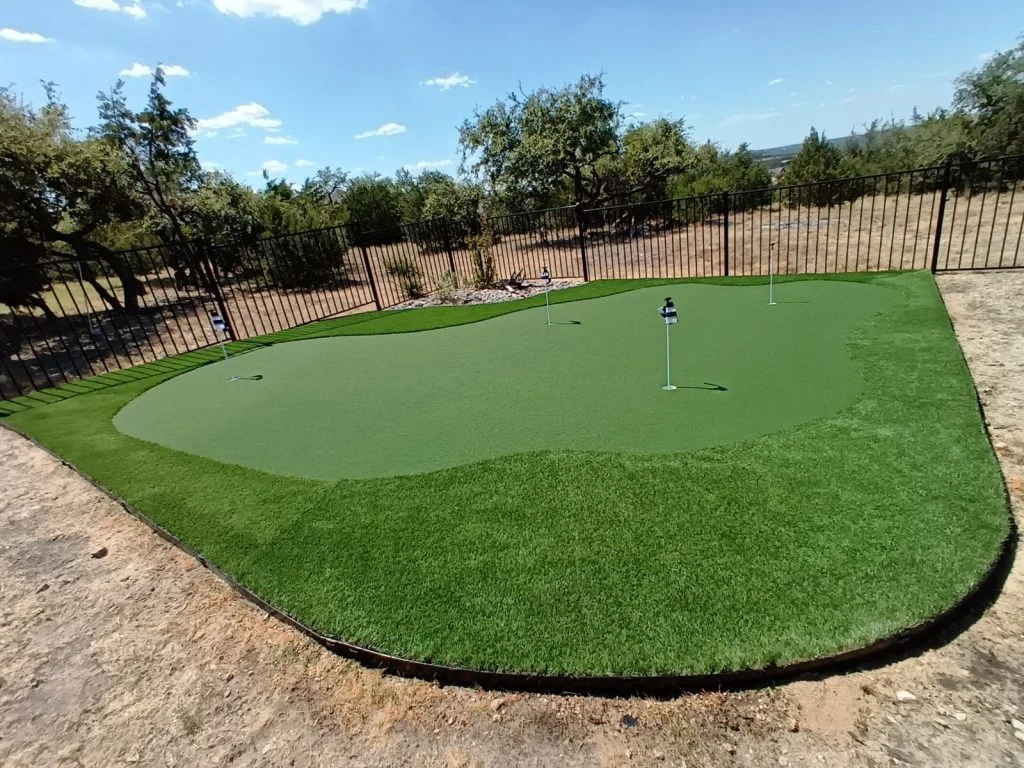 Putting green installed along a black metal fence. It has a longer pile dark green turf along the outside, and shorter pile putting turf towards the inside, with four holes indicated with flags.