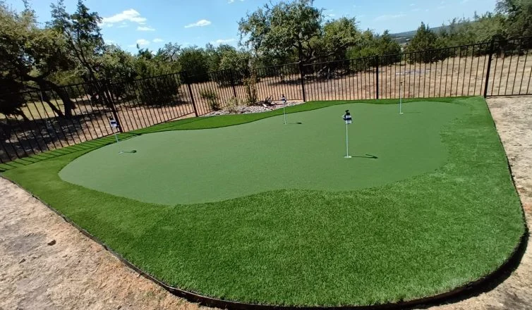 Putting green installed along a black metal fence. It has a longer pile dark green turf along the outside, and shorter pile putting turf towards the inside, with four holes indicated with flags.