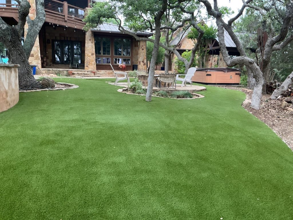 A completed artificial lawn installation in a residential backyard with several natural trees, an above-ground hot tub, and a stone seating area with a fire pit.