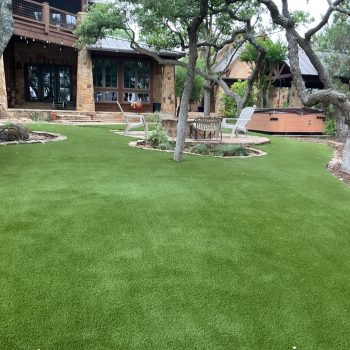 A completed artificial lawn installation in a residential backyard with several natural trees, an above-ground hot tub, and a stone seating area with a fire pit.