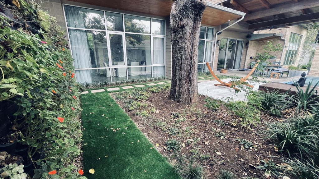 Photo depicting the need for artificial turf care in the yard of a house with a small patch of artificial turf next to the trunk of a large tree in real dirt to the right, and real shrubbery to the left. There are fallen leaves and other debris on the turf and the surrounding area.