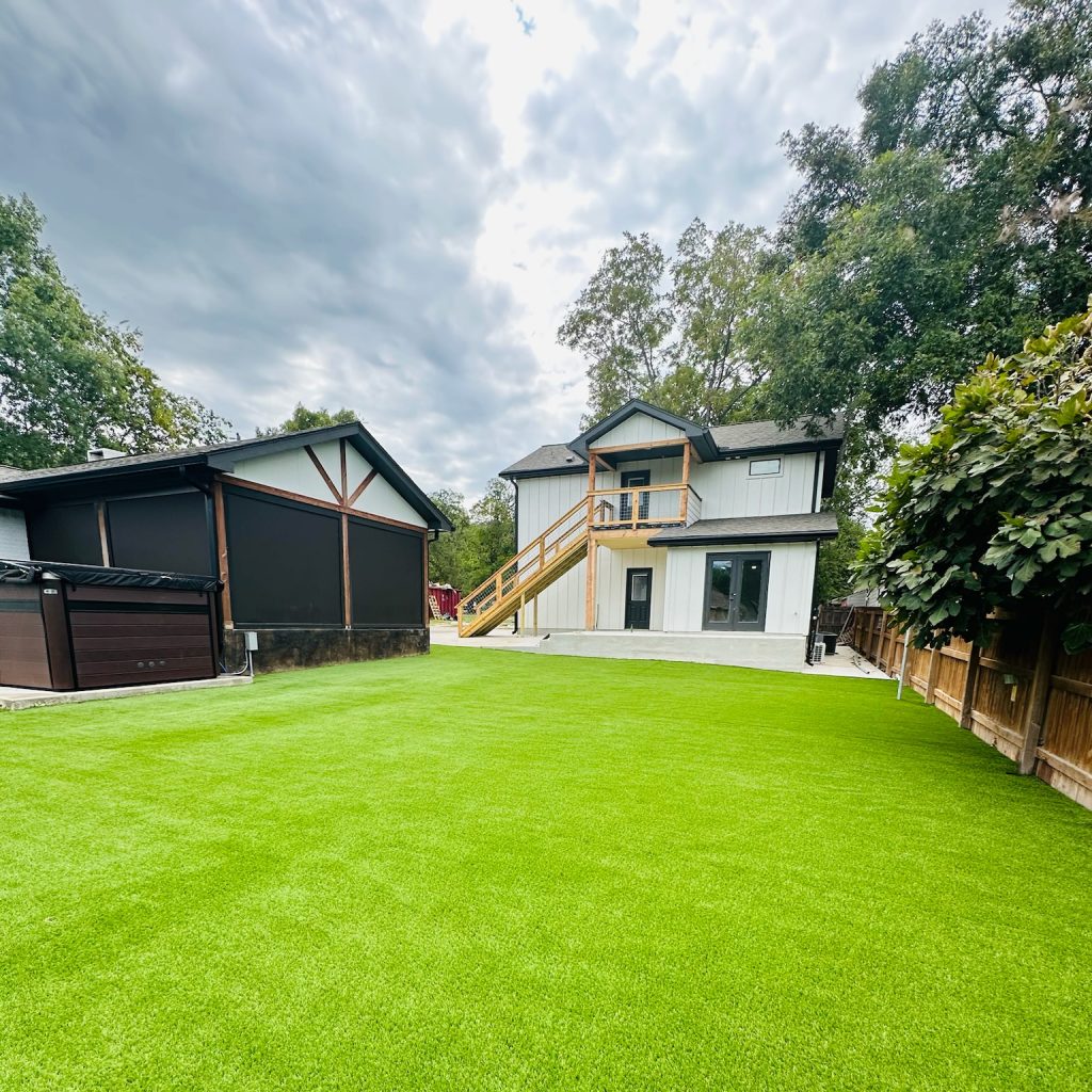 Backyard with artificial turf facing a white house with external stairs from the second floor to the ground, and a black shed to the left.