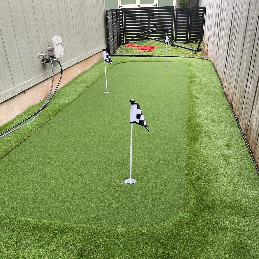 Small custom backyard putting green on the side of a house. It has three holes marked by black and white checkered flags.