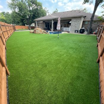 Turf installed in a long, narrow backyard, enclosed by a solid wood fence with string lights hanging along the perimeter. The house, a pool area, and a closed umbrella are in the distance.