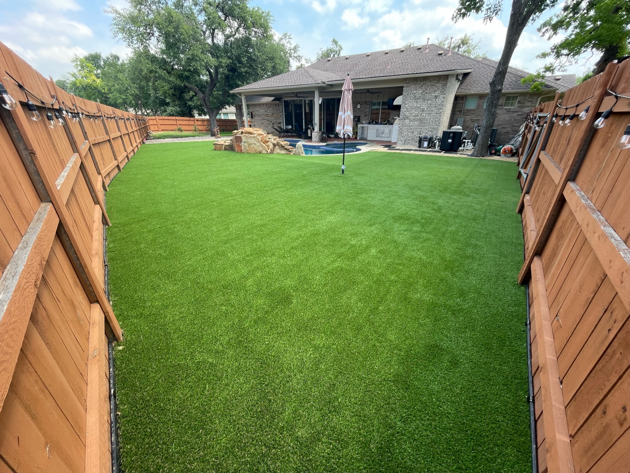 Residential Backyard Turf Install with Fence and Pool Turf installed in a long, narrow backyard, enclosed by a solid wood fence with string lights hanging along the perimeter. The house, a pool area, and a closed umbrella are in the distance.