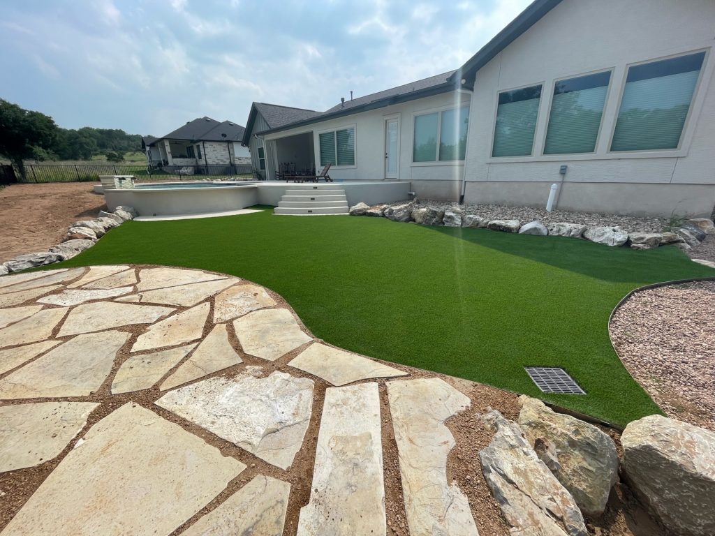 Professional artificial turf installation in Texas, showing a backyard with a stone patio and deck with a swimming pool, with turf installed in between