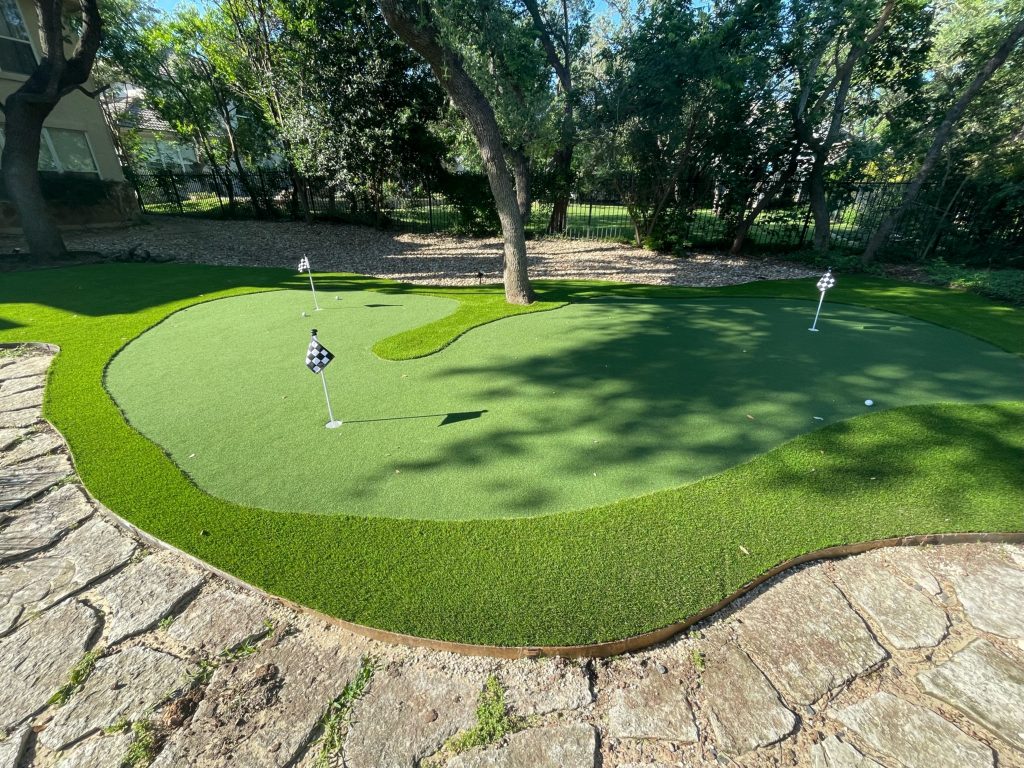 Custom backyard artificial turf putting green with three holes indicated with black and white checkered flags, shaped around a small tree in the center.