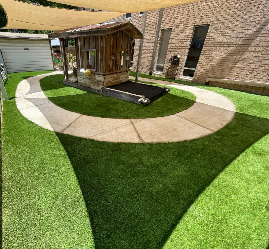 Does artificial turf get hot: an artificial turf play area with a curved concrete path and a wooden playhouse, shaded overhead by tan fabric shade sails. Does Artificial Turf Get Hot? The Honest Answer, Plus How Turf Handles Texas Weather. Lone Star Turf Texas.