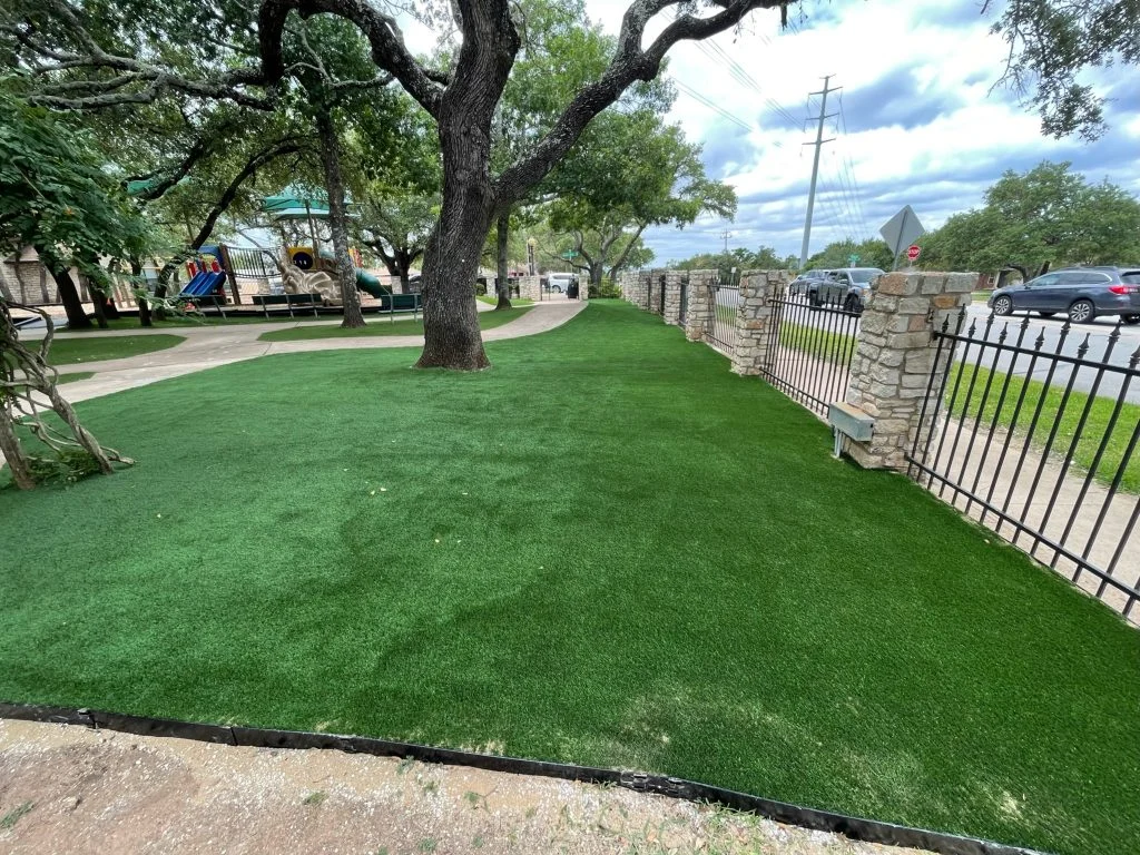 Artificial turf installed outside a commercial property on a main road, with stone columns separating sections of black metal fencing, and children's playground equipment in the distance.
