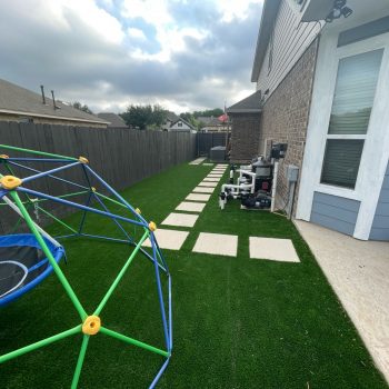 A kid's jungle gym and other toys on residential artificial grass installed along the side yard of a home with pavers leading to the door.