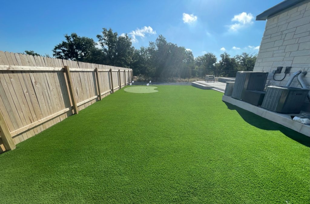 Backyard showing what you can get for the artificial turf cost in San Antonio: a large space with a small putting green along the side of a house and expanding into the backyard with a patio.