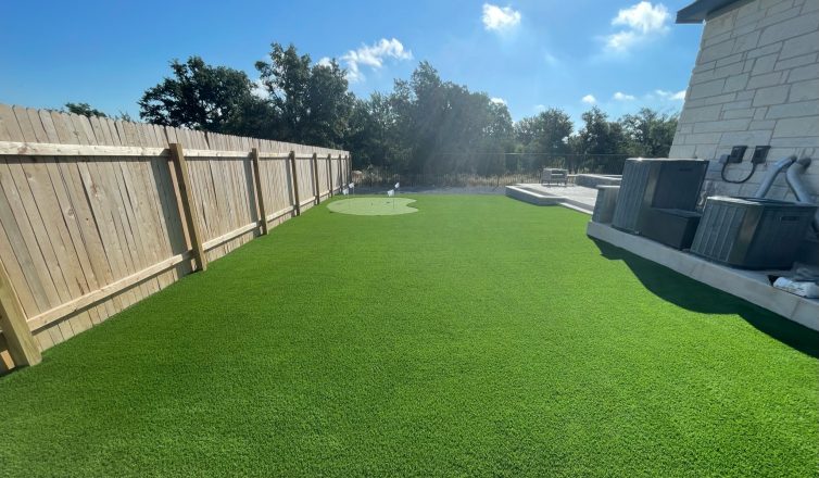 Backyard showing what you can get for the artificial turf cost in San Antonio: a large space with a small putting green along the side of a house and expanding into the backyard with a patio.