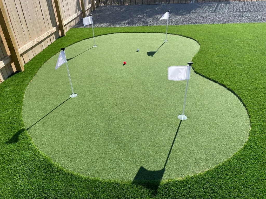 A backyard putting green with four holes marked by white flags. A white golf ball and a red golf ball are on the green.