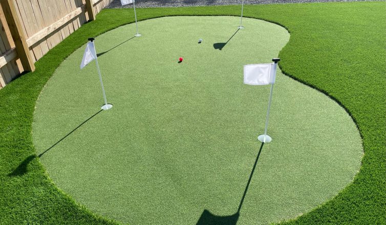 A backyard putting green with four holes marked by white flags. A white golf ball and a red golf ball are on the green.