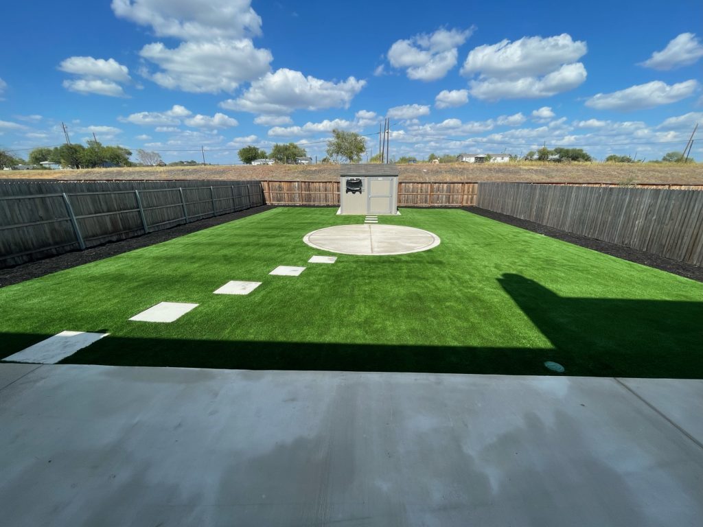 Photo of a backyard with artificial turf installed, featuring a walkway with stone pavers leading to a central concrete area, and additional pavers leading from the concrete area to a storage shed.