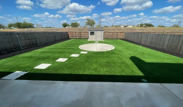 Photo of a backyard with artificial turf installed, featuring a walkway with stone pavers leading to a central concrete area, and additional pavers leading from the concrete area to a storage shed.