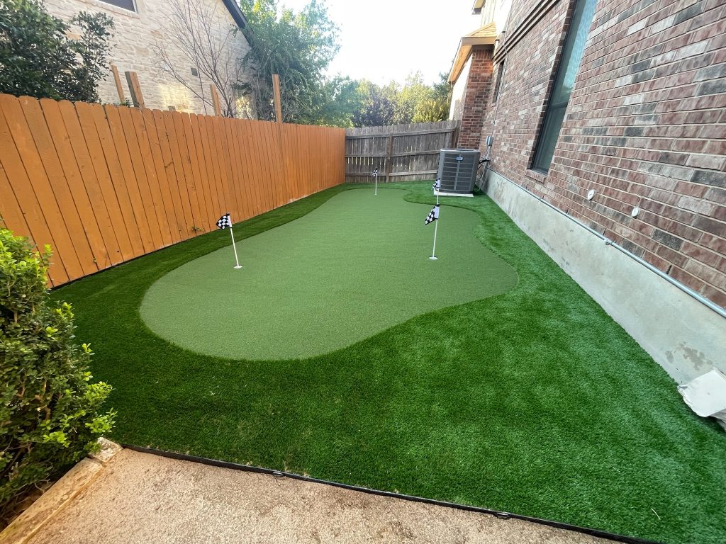 An artificial turf putting green along the side of a house, tucked between the wooden fence at the edge of the property and the brick exterior of the home. There are 3 holes marked with black and white checkered flags.