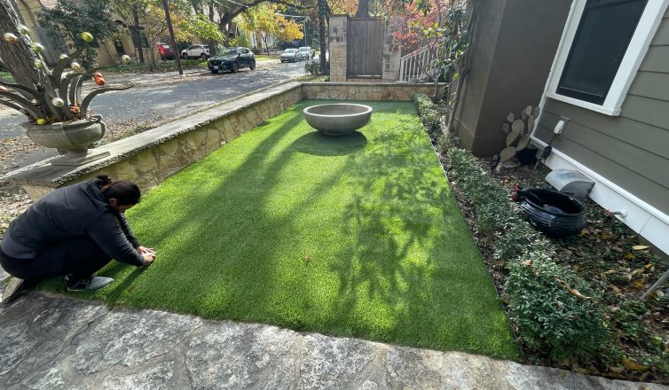 A small front yard area bordered by a stone pathway and low stone wall, with a professional turf installer making adjustments to the edge of the turf. A large decorative planter sits in the middle of the lawn, while surrounding greenery and neighborhood houses are visible in the background.