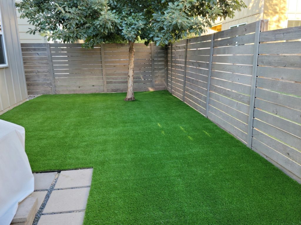  Artificial turf installed around a shade tree in a small Texas backyard with gray horizontal privacy fence and paver patio — How Much Does Artificial Turf Cost in Lakeway 2026 Guide by Lone Star Turf