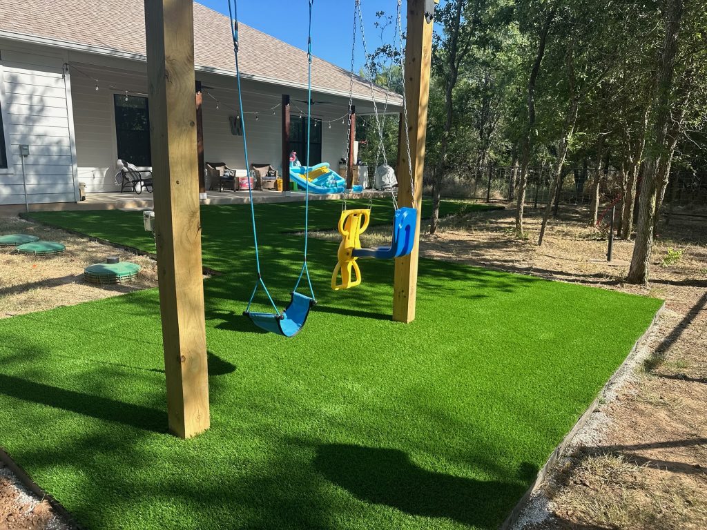 Does artificial turf get hot: a toddler swing set on an artificial turf play area shaded by nearby trees, with dry natural grass visible beyond the turf edge. Does Artificial Turf Get Hot? The Honest Answer, Plus How Turf Handles Texas Weather. Lone Star Turf Texas.
