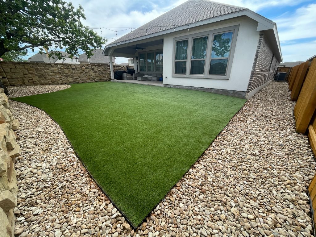 A backyard artificial lawn installation behind a white and brick house with a covered back porch. There is a wide border of large stone gravel between the artificial turf and the yard's fencing, which is wood on one side and stone on the other.