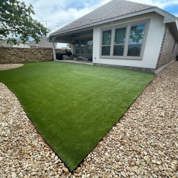 A backyard artificial lawn installation behind a white and brick house with a covered back porch. There is a wide border of large stone gravel between the artificial turf and the yard's fencing, which is wood on one side and stone on the other.