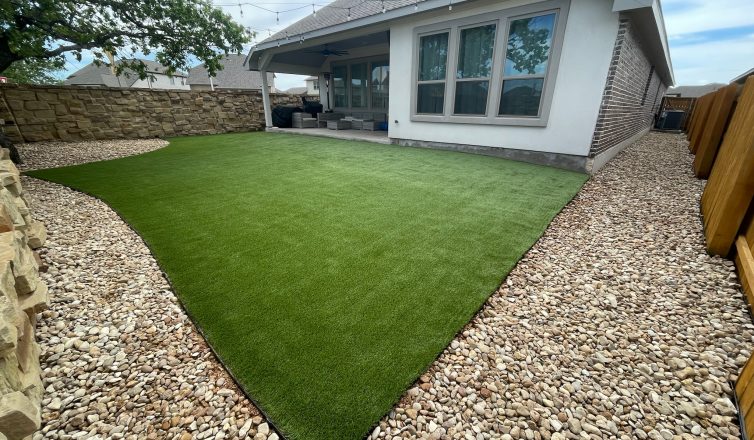 A backyard artificial lawn installation behind a white and brick house with a covered back porch. There is a wide border of large stone gravel between the artificial turf and the yard's fencing, which is wood on one side and stone on the other.
