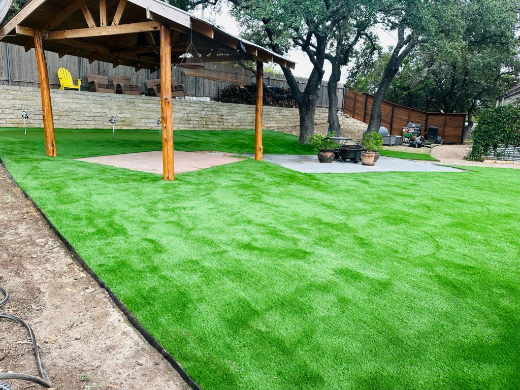 A spacious backyard with freshly installed artificial turf covering the ground. A wooden pergola stands over a paved square area in the center, with additional paved sections nearby. Potted plants and outdoor furniture are visible, along with a stone retaining wall in the background and a wooden fence lined with trees.