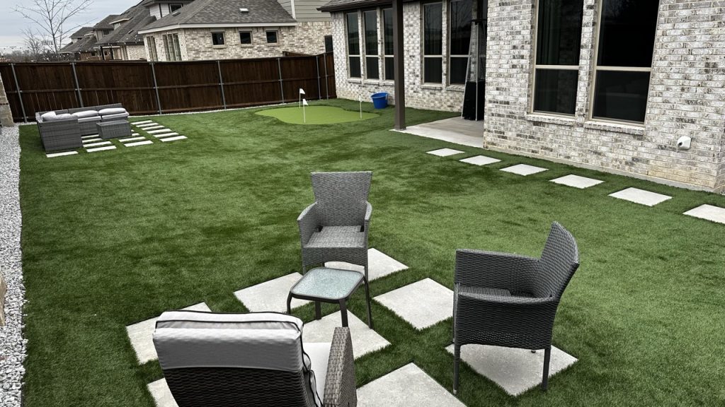 Backyard with multiple types of artificial turf. There are two seating areas on the main part of the turf, one in the foreground and one in the background. Both are on top of a grid of 9 paver stones. In the background to the right is a putting green in a lighter shade of turf. 