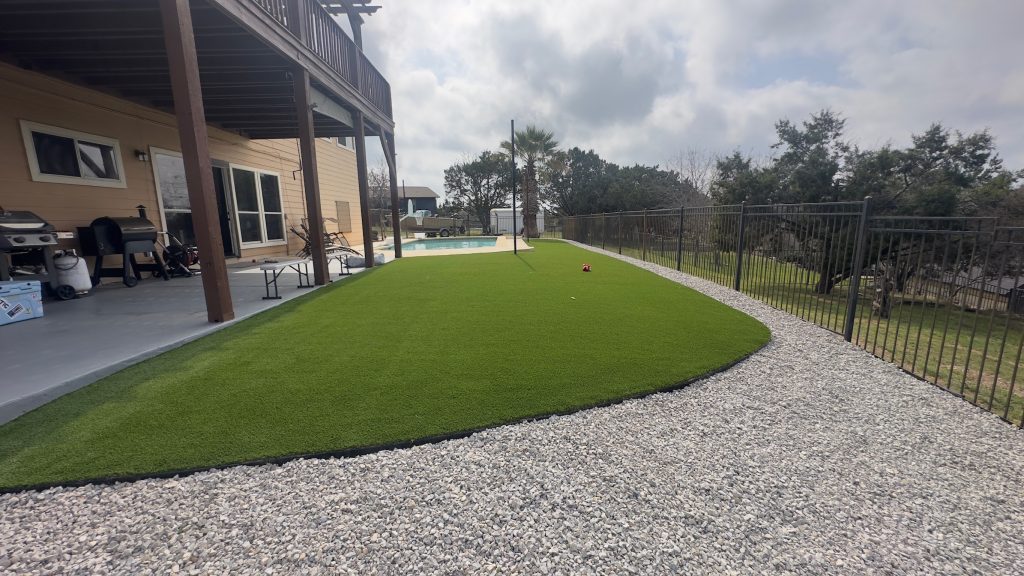 Artificial turf and decorative white stone gravel in a backyard area with a covered back porch on the left and a metal fence on the right.
