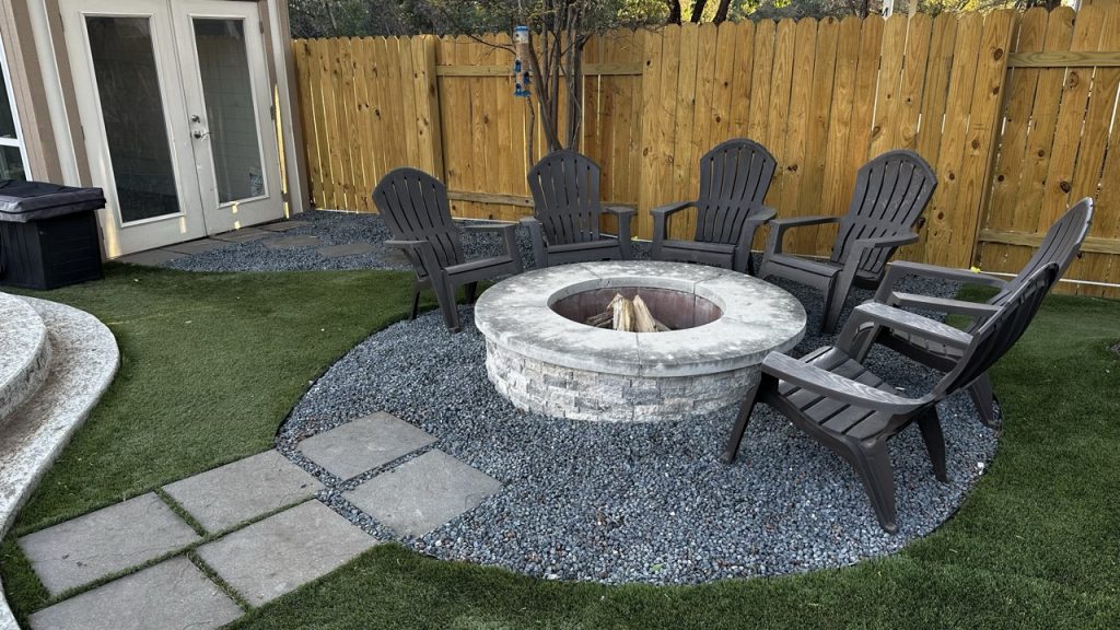 Artificial turf installation around a fire pit buffered with stone gravel. There are 6 Adirondack style chairs around the pit and stone pavers leading away from it.