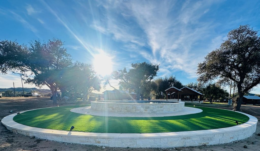 Does artificial turf get hot: a circular artificial turf lawn ringed by a limestone retaining wall around a raised stone pool, with live oak shadows falling across the turf in direct sun. Does Artificial Turf Get Hot? The Honest Answer, Plus How Turf Handles Texas Weather. Lone Star Turf Texas.
