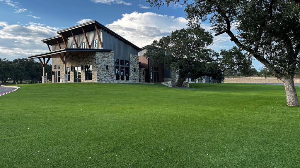 Photo of Nickel Rock HQ from the front, showcasing the vast artificial turf lawn with several natural old-growth trees and their brand-new building with a stone exterior on the lower level and metal siding on the upper level.