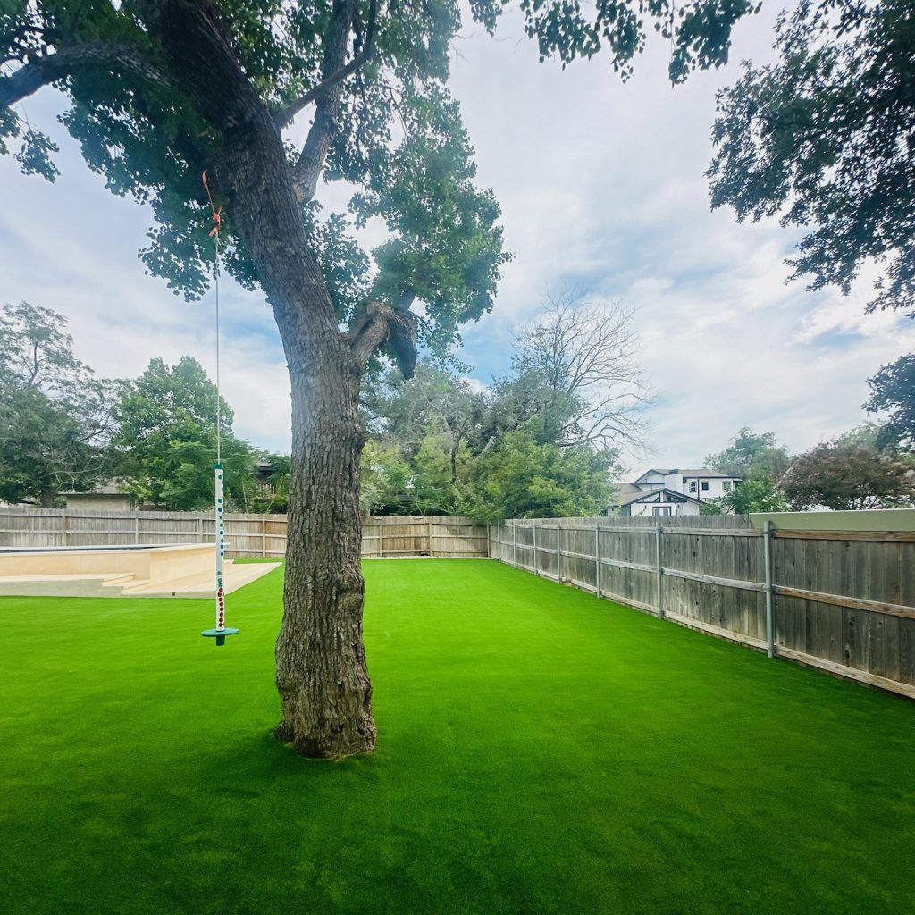 Artificial turf in a backyard, with a swing on a large tree in the middle.