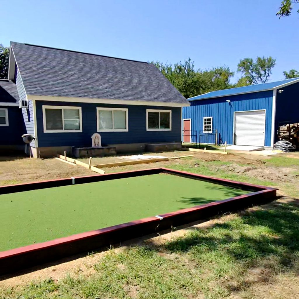 Half of a regulation sized bocce ball court, with two by fours installed as a border. Behind the court is the back of a house with blue siding.