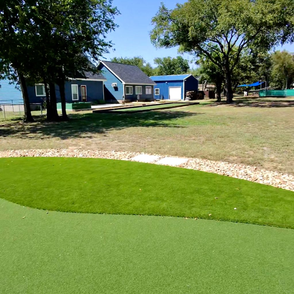 Photo taken while standing in the middle of a large putting green, facing a house with blue siding at the other end of the yard. The green has a stone gravel border around it and two square stone pavers are visible in part of the border for easy access.