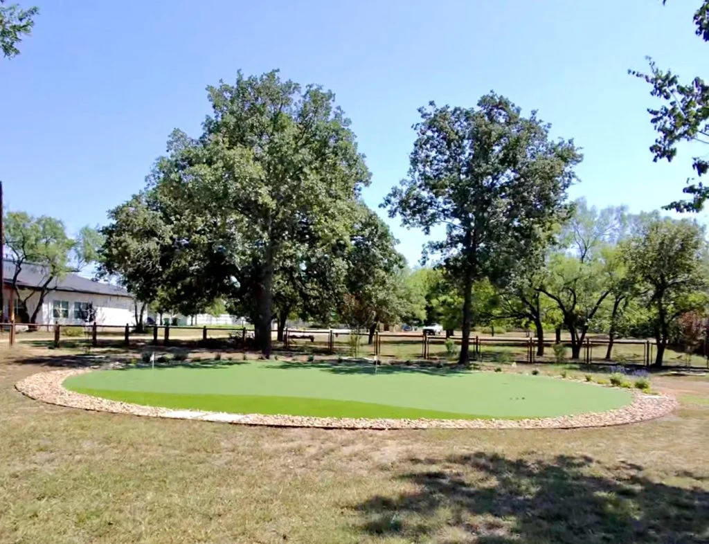 A large backyard putting green with a stone gravel border and two large trees behind it, providing partial shade.