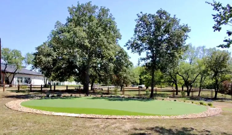 A large backyard putting green with a stone gravel border and two large trees behind it, providing partial shade.