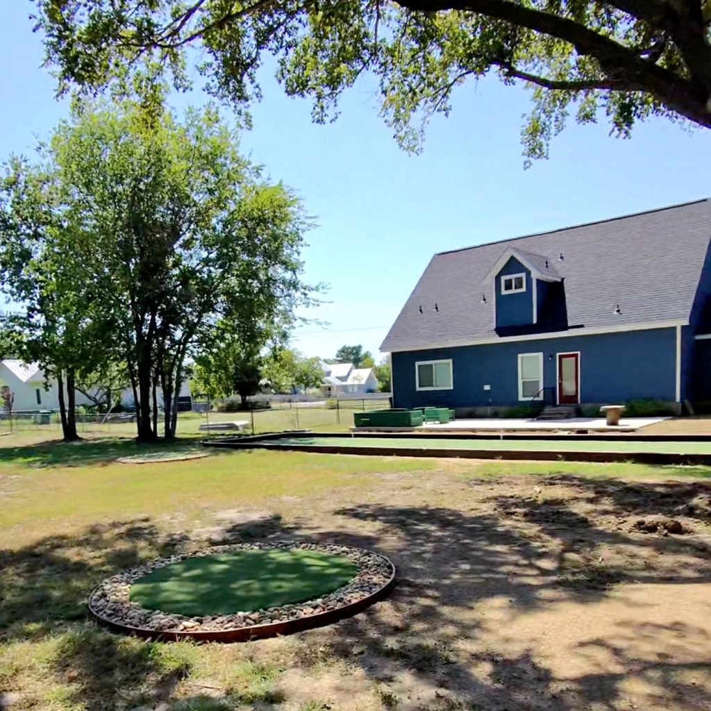 Backyard of a blue house with a small golf chipping mat with a stone gravel border in the foreground and a bocce ball court in the background.