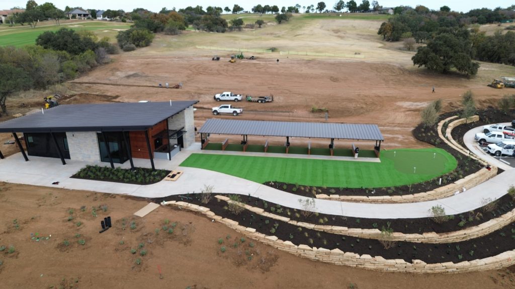 Overhead photo from a drone of a fresh artificial turf installation with a putting green, creating a bright pop of green in the middle of a large area of undeveloped dirt.