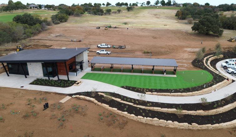 Overhead photo from a drone of a fresh artificial turf installation with a putting green, creating a bright pop of green in the middle of a large area of undeveloped dirt.