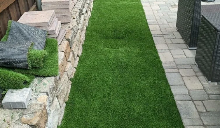Side yard with artificial turf installed and additional installation materials to the left side on a stone half-wall.